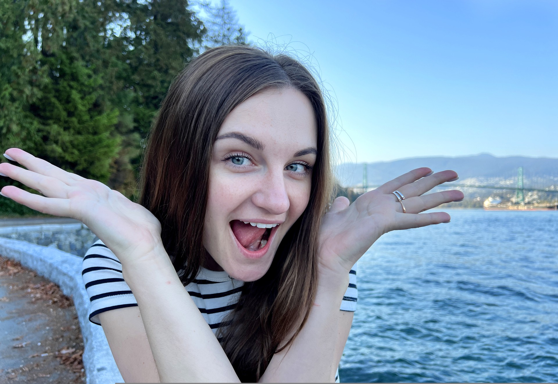 young woman 25 years old laughs, shows her hands to the side and opens her mouth saying WoW She sits Stanley Park Vancouver Canada Travel advertising for travel agencies Striped nautical t-shirt